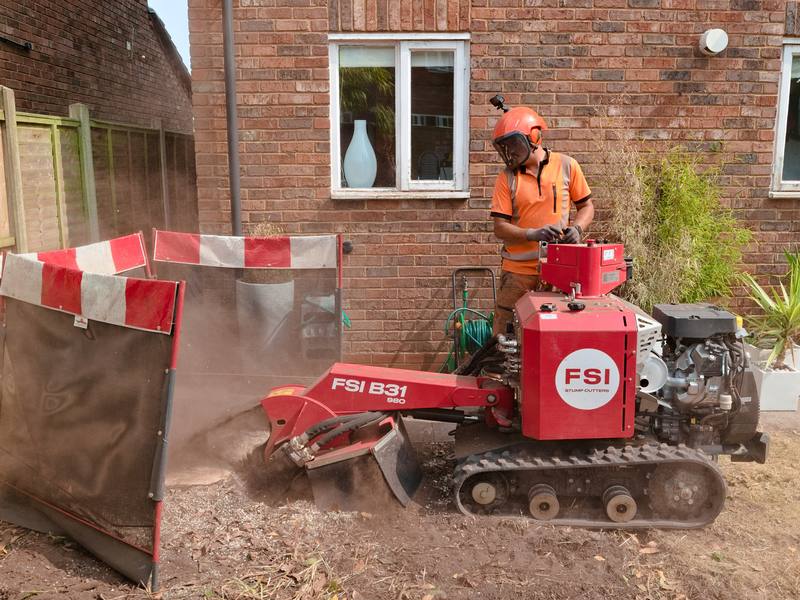 Tracked stump grinder breaking a stump down below ground level beside a house, with guard screens up